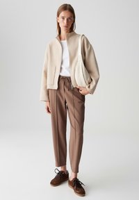 Femme portant une veste beige zippée, une chemise blanche, un pantalon marron cintré et des baskets marron, portant un sac à épaule beige, se tenant sur un fond gris.