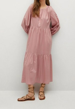 Femme portant une robe midi à manches longues rose avec une taille froncée et des sandales plates à lacets de couleur beige, sur un fond blanc uni.