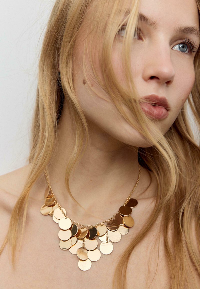 Blonde vrouw met blauwe ogen die een gouden ketting draagt bestaande uit meerdere ronde, glanzende schijven, met haar dat gedeeltelijk haar gezicht bedekt.