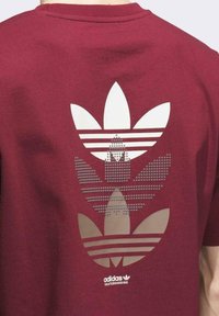 Camiseta de algodón marrón con un gran gráfico texturizado de tres logotipos de trébol superpuestos en gris claro en la parte posterior.
