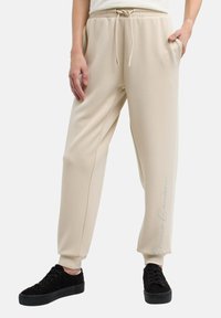 Beige sweatpants med elastisk midjeband och snöre, med sidofickor och en subtil texttryck på benet. Tillsammans med svarta sneakers.