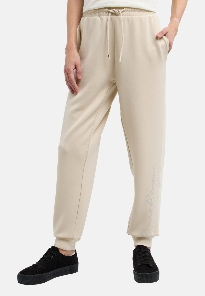 Beige sweatpants med elastisk midjeband och snöre, med sidofickor och en subtil texttryck på benet. Tillsammans med svarta sneakers.