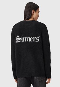 Osoba z długimi ciemnymi włosami, ubrana w czarny, puszysty sweter z białym, gotyckim napisem "Sinners" na plecach, w połączeniu z ciemnymi dżinsami.