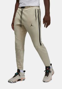 Beiga träningsbyxor med svarta sidostreck och en Jumpman-logga. Har en dragsko i midjan och en avsmalnande passform, ihop med lätta sneakers.