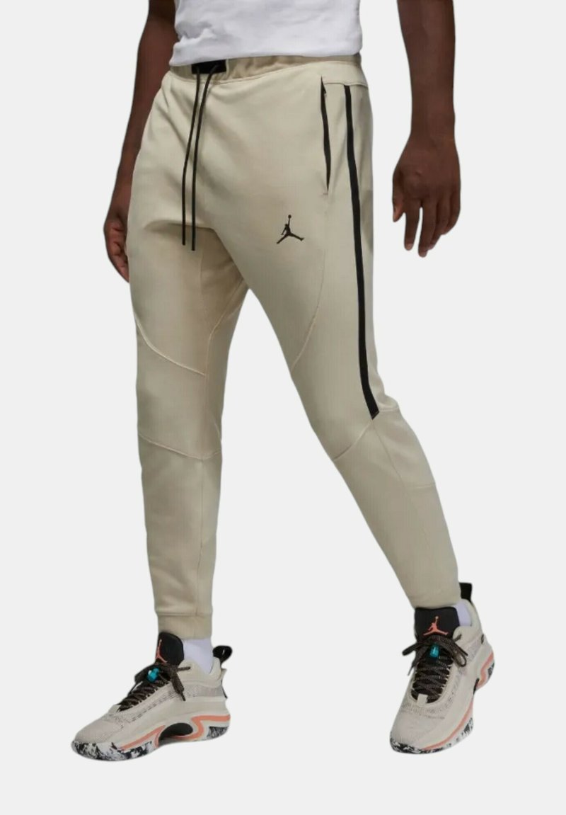 Beiga träningsbyxor med svarta sidostreck och en Jumpman-logga. Har en dragsko i midjan och en avsmalnande passform, ihop med lätta sneakers.