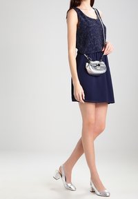 Marinblå ärmlös klänning med en texturerad pärlbeklädd överdel, tillsammans med en metallic silver handväska och silverblockklackar.