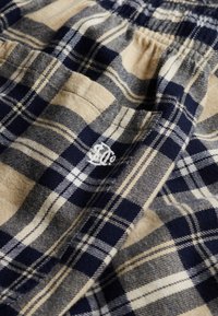 Gros plan sur un tissu à carreaux bleu marine et beige avec des coutures visibles et un logo blanc brodé sur un matériau doux et texturé.