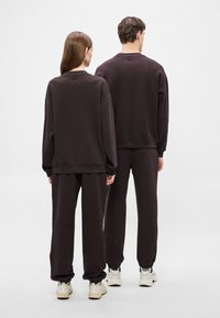 Σκούρα καφέ φούτερ και παντελόνια από απαλό ύφασμα· άνετη εφαρμογή με ριμπ μανσέτες, φορεμένα με λευκά αθλητικά παπούτσια, από πίσω όψη.