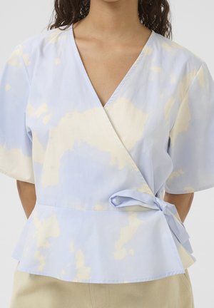 Kobieta w pastelowo niebieskiej i kremowej bluzce z krótkim rękawem, wiązanej z boku, w połączeniu z jasnymi spodniami.