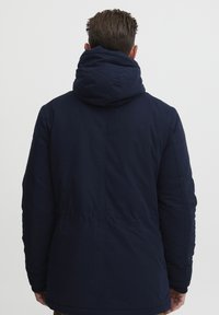 Navy blue puffer jacket with a large, padded hood. Smooth texture and fitted silhouette with elastic cuffs and a seam at the waist.