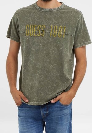 Homme portant un t-shirt vert olive délavé avec l'inscription brodée "GUESS 1981" en jaune, associé à un jean bleu, les mains dans les poches.