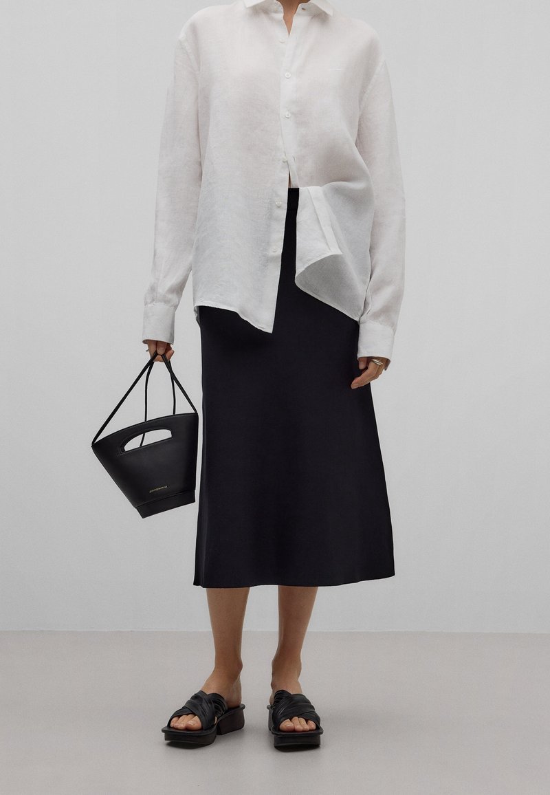 Personne portant une chemise blanche ample boutonnée, une jupe midi noire, des sandales à plateforme noires et tenant un petit sac à main noir.