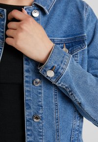 Jeansjacka med blåtvätt, silverfärgade tryckknappar och kontrasterande sömmar. Ärmarna har sömdetaljer och justerbara knäppningar.