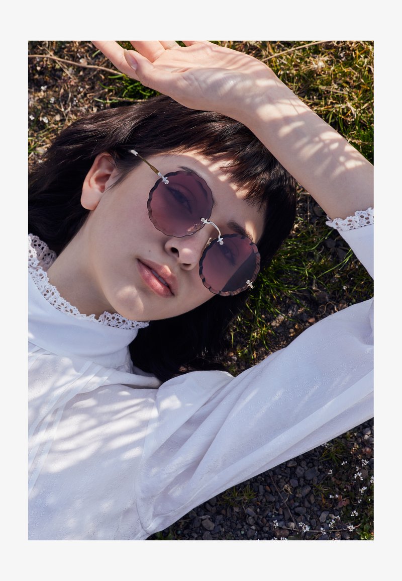 Mladić s tamnom kosom leži na travi i šljunku, nosi okrugle ljubičaste sunčane naočale i bijelu majicu s čipkanim ovratnikom, zastire oči podignutom rukom.