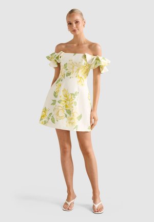 Žena stojaca v bielych šatách s odhalenými ramenami, žltým kvetinovým vzorom a volánmi na rukávoch, doplnená bielymi sandálmi s otvorenou špičkou.