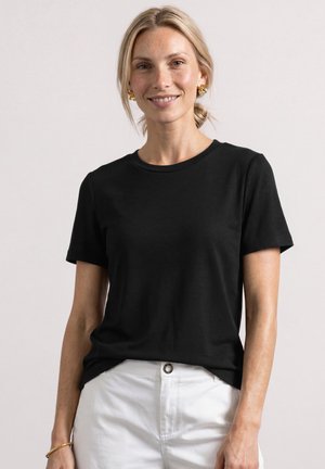 Lyshåret kvinde smiler, iført sort kortærmet skjorte, hvide bukser, guldbelagte øreringe og armbånd, stående mod en ensfarvet lys baggrund.