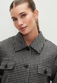 Giacca pied de poule grigia e nera con colletto, due tasche sul petto, bottoni neri e finitura del tessuto strutturata.