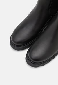 Bottines en cuir noir avec une finition lisse, bout rond et panneaux latéraux élastiques ; dotées d'une semelle en caoutchouc noir texturé.