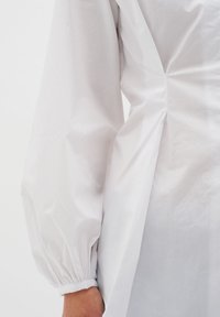 Biała bluzka z długim rękawem, z marszczonym mankietem i bocznymi zaszewkami, wykonana z gładkiego, lekkiego materiału. Minimalistyczny design.