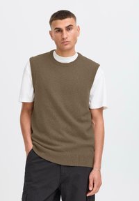 Suéter sin mangas de punto marrón sobre una camiseta blanca, con cuello redondo y bordes acanalados, de corte holgado y textura suave.
