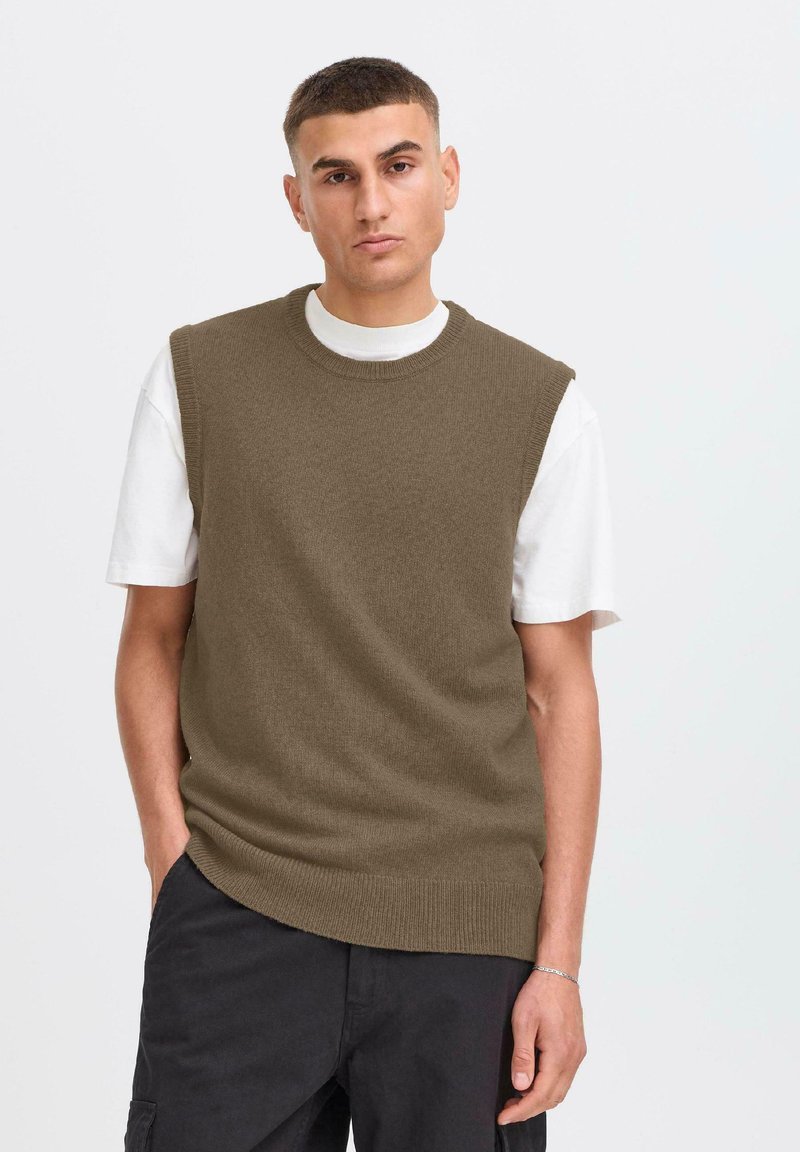 Suéter sin mangas de punto marrón sobre una camiseta blanca, con cuello redondo y bordes acanalados, de corte holgado y textura suave.