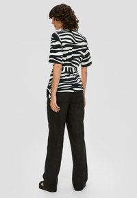 Vrouw met krullend haar, gekleed in een shirt met zebraprint met korte mouwen, zwarte wijde broek en zwarte sandalen, staand tegen een grijze achtergrond.