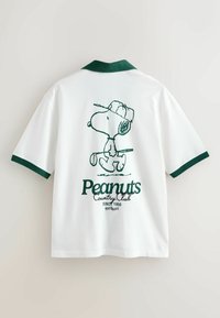 Hvid poloshirt med grøn krave og ærmekanter, med et grønt omrids af Snoopy, der spiller golf, og teksten "Peanuts Country Club" på ryggen.