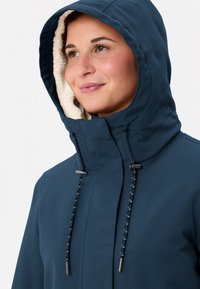 Vrouw in een marineblauwe hoodie met een fleece-voering op de capuchon, die iets omhoog kijkt met een zachte glimlach op een witte achtergrond.