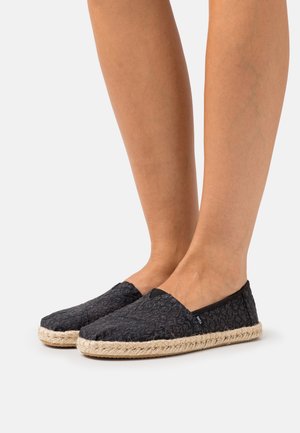 Benen in zwarte, gestructureerde espadrille instapschoenen met gevlochten jute zolen op een lichte achtergrond.