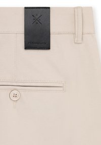 Beżowe spodnie z czarną skórzaną metką z wytłoczonym logo, zapięciem na guzik oraz tylną kieszenią z poziomym otwarciem.