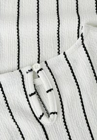 Tissu texturé blanc avec des rayures noires. Dispose d'une fermeture à boutons et d'un col arrondi. Le matériau semble léger et respirant.
