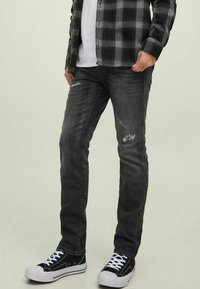 Jeans ajustados de mezclilla negra con detalles desgastados, combinados con zapatillas en blanco y negro y una camisa de franela a cuadros negra sobre una camiseta blanca.