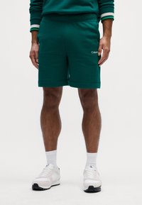 Grønne bomuldsblandingsshorts med elastisk talje, sidelommer og logodetalje. Parret med hvide sneakers og sokker.