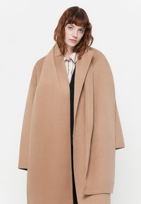 Beige wollen jas met een brede, open voorkant en oversized mouwen. De stof heeft een gladde textuur, wat zorgt voor een minimalistisch ontwerp.
