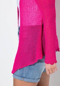 Ljusröd öppenstickad cardigan med vågiga kanter, lagd över en vit topp och parat med rullade denimshorts. Strukturerad tyg.