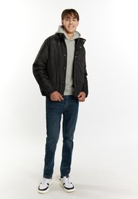 Jachetă neagră umplută cu capse, hanorac gri dedesubt, blugi albaștri și adidași albi cu accente negre. Modelul stă în picioare pe un fundal alb.