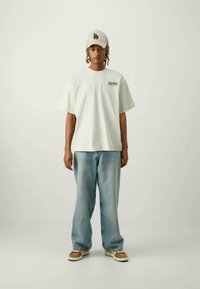 Bílé oversized tričko s potiskem "RESONANCE", světle modré baggy džíny, béžové tenisky a světle šedá kšiltovka s logem "LA".