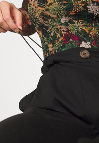 Pantalon noir taille haute avec fermeture à bouton et détail cordon, associé à un haut à motifs floraux multicolores. Texture lisse.