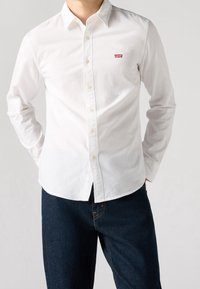 Camicia bianca button-up in cotone, con colletto classico, maniche lunghe e un piccolo logo rosso di Levi's sul petto. Abbinata a jeans scuri.