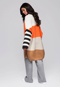 Długi sweter wykonany na drutach w paski o kolorach pomarańczowym, kremowym i brązowym, z czarno-białymi paskami na rękawach. Luźny krój z prążkowanymi mankietami i dolnym brzegiem.