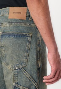 Denim jeans in light blue with a rough texture, featuring a leather patch labeled "EIGHTYFIVE," and unique pocket design with metal accents.