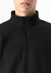 Hombre vestido con un pulóver de forro polar negro texturizado, con un alto cuello y una media cremallera en la parte delantera, sobre un fondo blanco.