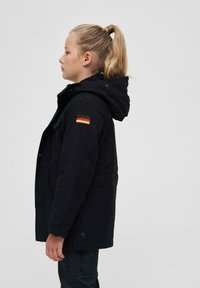 Črna kapucar jakna z ovratnikom obloženim s flisom, z oznako nemske zastave na levem rokavu. Tkanina ima teksturiran zaključek.