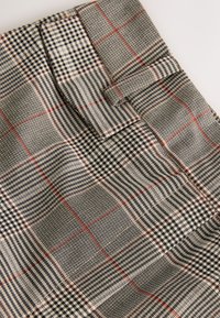 Tessuto a quadri grigio e nero con motivi a griglia variabili e accenti rossi. Texture liscia con un design strutturato e dettaglio piegato.