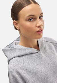 Sudadera con capucha de color gris claro, fabricada con tejido suave, que presenta un corte holgado y detalles de costura mínimos. La lleva puesta un modelo con el cabello recogido.