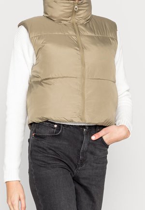 Beżowa pikowana kamizelka z wysokim kołnierzem, zamkiem błyskawicznym z przodu i pikowaną fakturą. Noszona na białej, prążkowanej bluzce z długim rękawem i czarnych dżinsach.