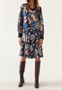 Blumendruckkleid mit langen Ärmeln, V-Ausschnitt, mehrlagigem Rüschensaum und in dunklem Blau, Pink und Grün; kombiniert mit kniebedeckenden braunen Stiefeln.