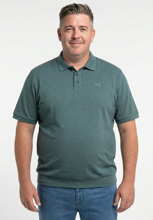 Hombre con cabello corto y gris que lleva una camiseta tipo polo verde oscuro y jeans azules, de pie contra un fondo blanco.