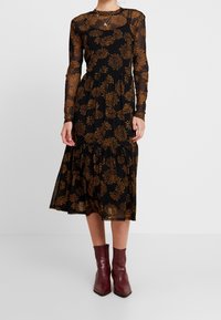 Kvinna som bär en svart midi-klänning med genomskinliga långa ärmar och bruna blommönster, matchad med mörkröda ankelskor.