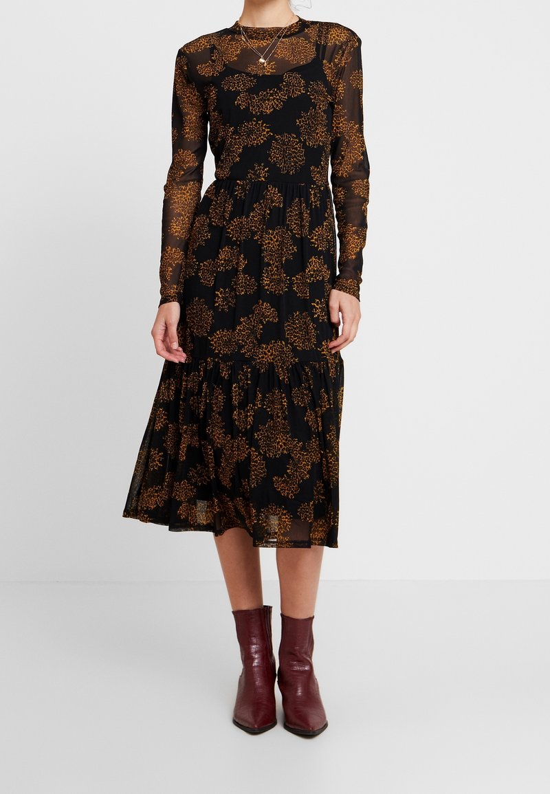 Kvinna som bär en svart midi-klänning med genomskinliga långa ärmar och bruna blommönster, matchad med mörkröda ankelskor.